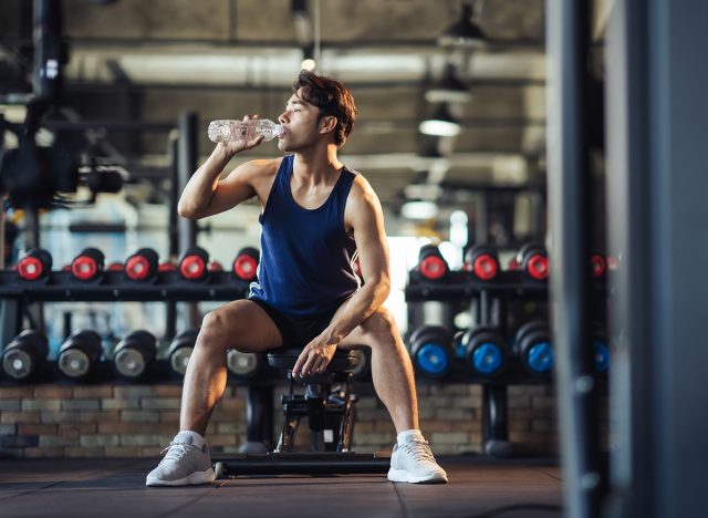 Exhausted man sitting drinking the water after weight training in fitness gym. Health care and workout. Asian man handsome replenishing water balance after workout.