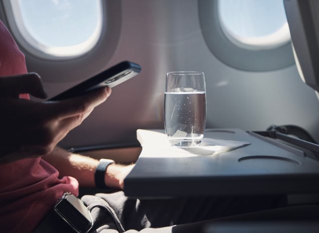 Glass of water on table of seat in airplane. Passenger is using phone during flight.