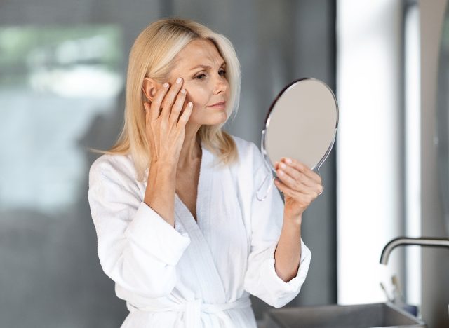 Skin Aging. Attractive Mature Woman Holding Mirror And Looking At Her Wrinkles Near Eyes, Beautiful Senior Female Wearing Bathrobe Checking Face, Noticing Age Changes, Standing In Bathroom Interior