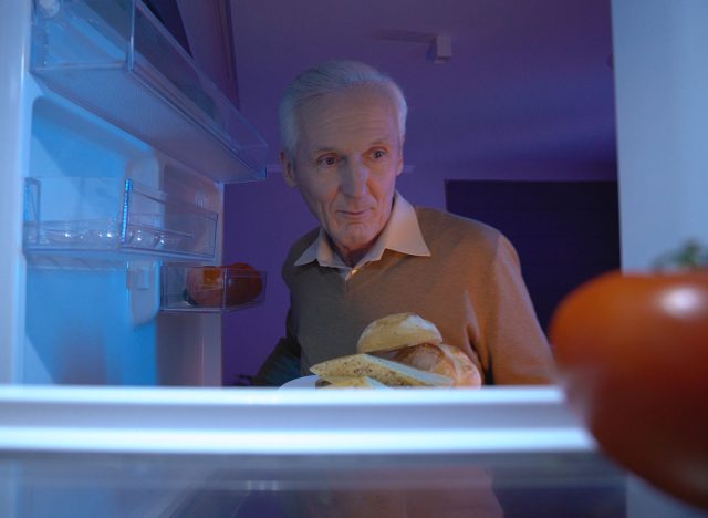 A hungry senior man is taking a plate with meats and bread from the fridge, overeating late at night