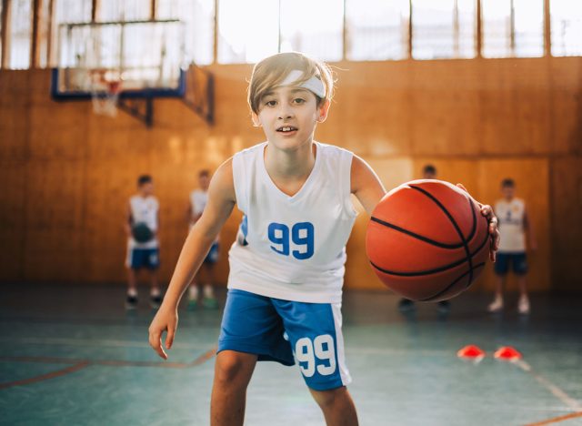 a junior athlete dribbling a ball towards camera and practicing at indoor court. There are his teammates in a blurry background. A young basketball player playing basket on court.