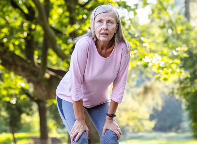 Tired senior gray-haired woman doing sports, jogging and walking in the park, standing with hands on knees and resting, breathing heavily.