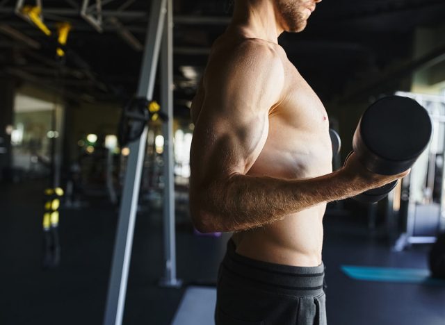 Shirtless man showcasing muscle definition while holding a dumbbell in a gym.