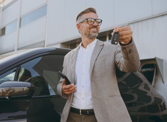 Adult man customer male buyer client wearing white shirt classic grey suit standing near black electric car while charging looking aside use mobile cell phone hold give keys outside. Business concept