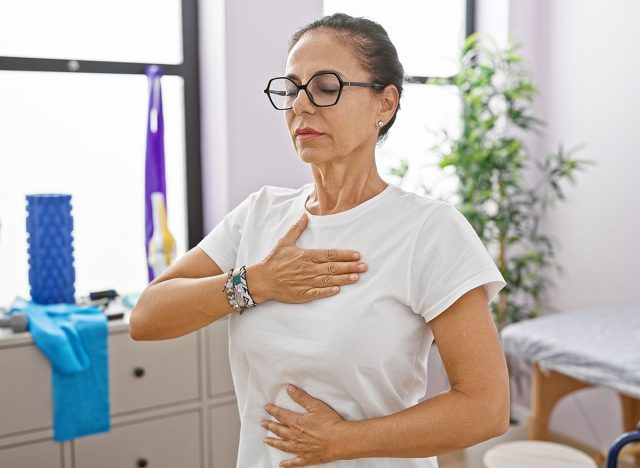 Hispanic senior woman with glasses and curly hair practicing deep breathing exercises indoors at a rehabilitation center.