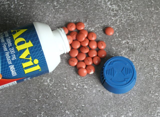 A white bottle of Advil, containing 200 mg ibuprofen tablets, is displayed with the cap removed and several round, orange tablets scattered on a gray, textured surface.