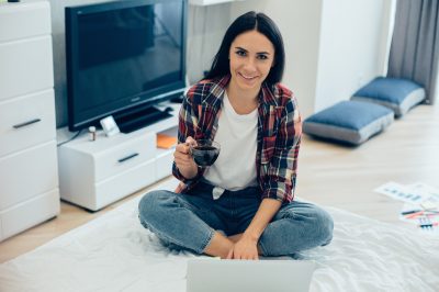 Positive smiling young woman sitting cross-legged on the bed in front of a laptop and holding a cup of coffee