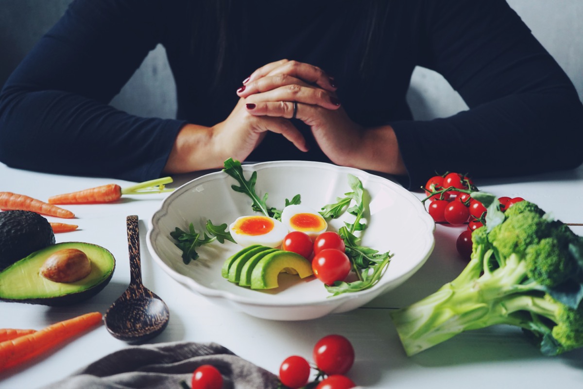 Weight loss plans for women theme white ceramic bowl contained healthy salad (slided avocados, boiled eggs, rocket leaves, tomatoes) on wooden table with blurred woman in black dress / selective focus