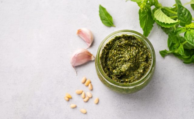 Traditional Italian pesto in a jar with green basil, pine nuts and garlic on a light background. A classic sauce for spaghetti or bruschetta. Delicious vegetarian homemade food. Top view