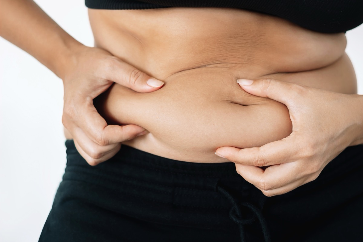 A cropped shot of a young woman in a black underwear holding herself by the fat on her stomach isolated on a white background. Overeating, overweight, excess weight, hormonal belly, rolls