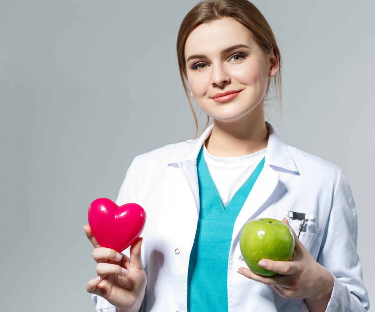 Beautiful smiling female doctor holding red heart and green apple in front of chest. Health life and wholesome food concept. Vegetarian lifestyle concept. Cardiology concept