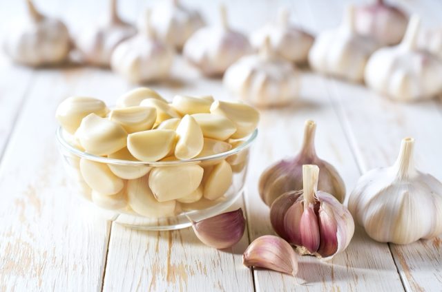 Garlic cloves in a glass bowl with peeled garlic on a white wooden table.