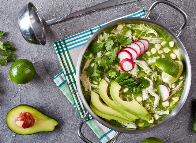 Chicken Pozole Verde in a pot - Mexican stew filled with shredded chicken and hominy in a warm green chile broth garnished with radish slices, fresh green coriander and a lime, close-up