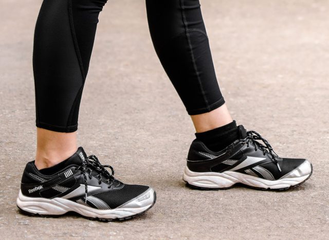 Attractive healthy woman in black gym tights and shoes, standing at ease with one foot forward. Walk, exercise, workout. Reebok shoes.