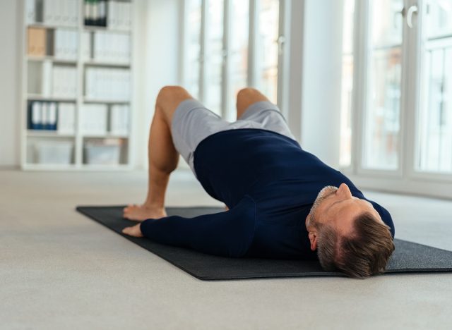 Man doing bridging exercise, lying on his back on black mat in empty office interior. Viewed from floor level from his head, glute bridge