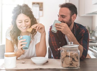 Engaged couple has breakfast together in their new home - young couple smiling while drinking and eating in the kitchen - warm filter on background