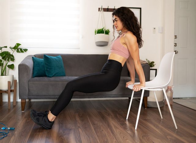 Attractive young woman exercising in her home. Sporty woman in her 20s doing tricep dips using a chair in the living room