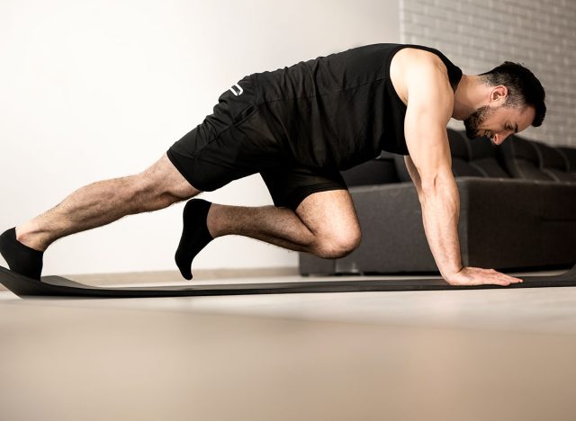 Man doing mountain climber exercises on black yoga mat. Morning workout. White modern living room on background. Plastic bottle of water. Hard workout. Training at home.