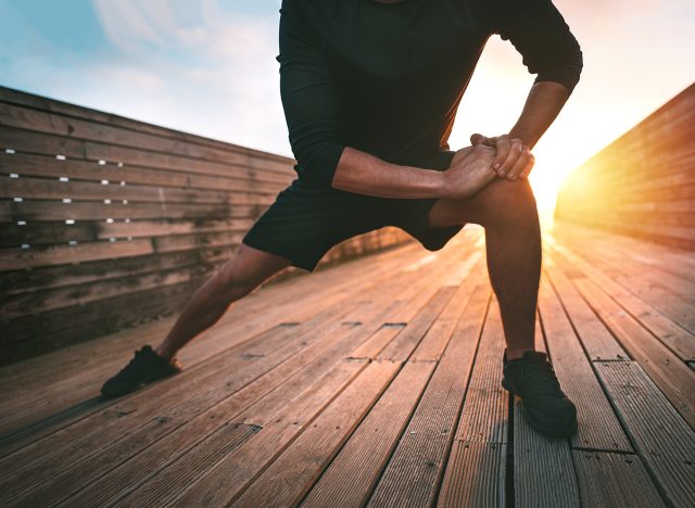 Sporty man stretching leg adductor muscles and warming up for training outdoors at sunset or sunrise. Athletic man in black sportswear doing fitness stretching exercises. Sport and healthy lifestyle