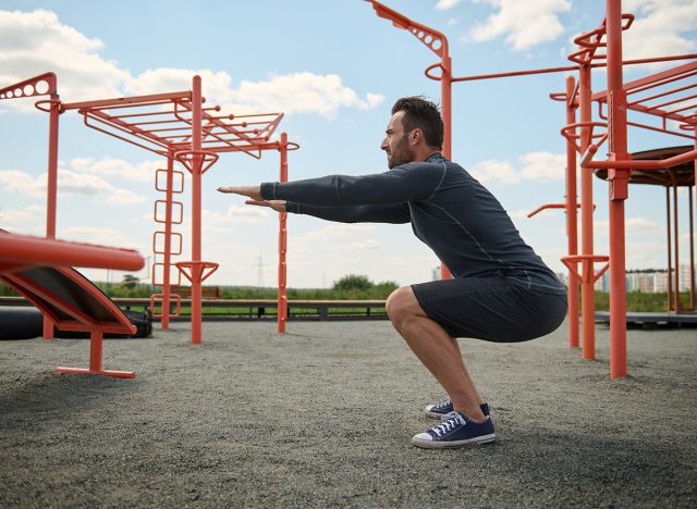 Active athlete in athletic uniform performing bodyweight squats during training on an outdoor sports field. Young bearded man doing sports on summer sportsground. Healthy active lifestyle concept