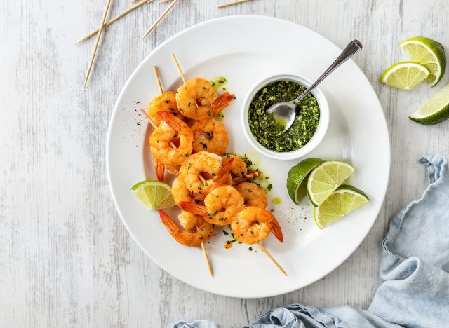 Shrimp skewers grilled, served with green homemade pesto and lime slices on white plate top view on wooden table