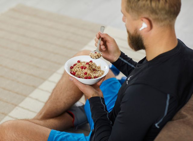 Athlete enjoying healthy meal rich in fiber, protein and vitamins. Fit young man sitting on floor in living room, relaxing after fitness workout, eating natural vegetarian granola, listening to music