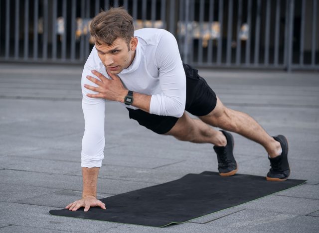 Focused man in sportswear doing shoulder tap in push or press ups exercise, standing in plank pose. Side view of confident and serious sportsman with smart watch training outdoor, work out on yoga mat