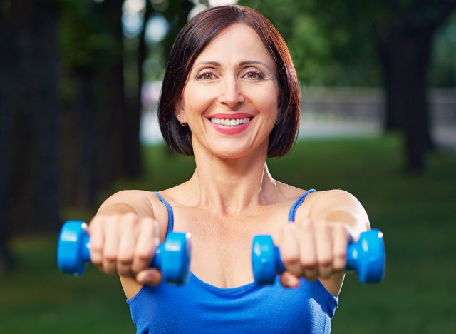 Portrait of cheerful aged woman in fitness wear exercising with dumbbells in park.