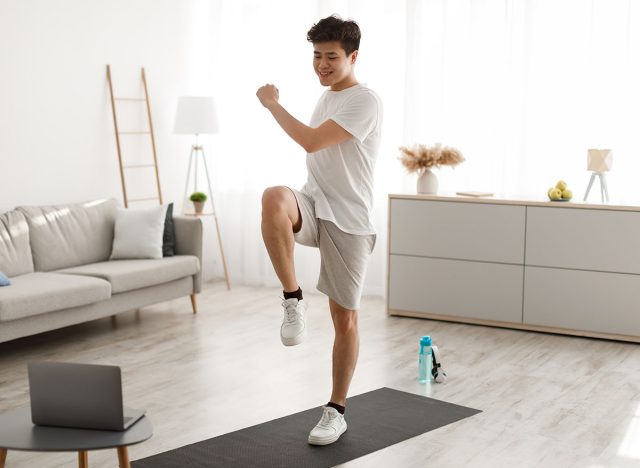 Online Workout Concept. Cheerful Smiling Asian Guy Training In Living Room, Doing Twist Knees To Elbow Exercise. Happy Man Standing And Lifting Leg Up, Using Laptop Watching Tutorial Lookign At Screen, single leg raise, 