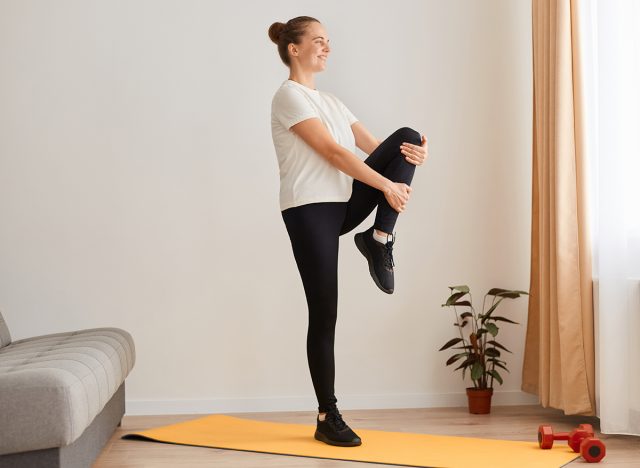 Side view of woman exercising by lifting the legs, knees up one by one, standing straight back and hold, relaxing yoga practice at home, wearing white t shirt and black leggins.