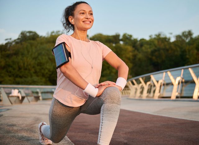 Beautiful middle aged sportswoman with earphones and smartphone holder enjoying morning workout at sunrise, stretching legs, doing lunges, squatting before jogging. Female athlete working out outdoor