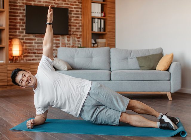 Happy asian middle aged man in sportswear doing side plank on floor mat in living room interior, copy space. Sport at home, workout, fitness and exercises, active lifestyle