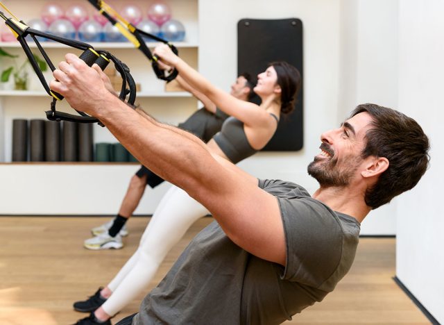 Side view of bearded male athlete smiling and doing low row exercise with TRX ropes during group suspension training in gym