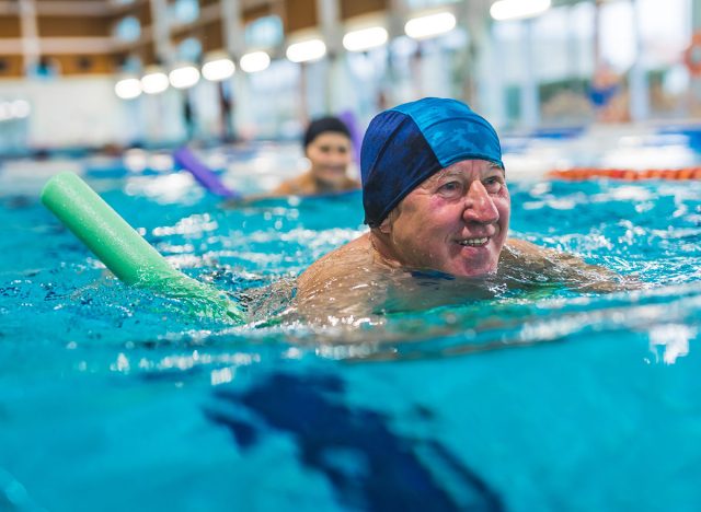 Portrait of smiling happy positive white senior adult man with blue swimming swim cap on his head using foam green pool noodle. Indoor shot. Sports area. High quality photo