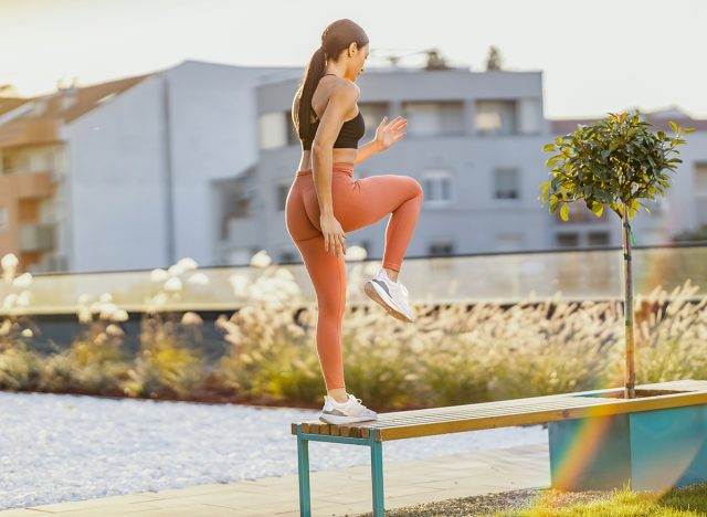 A fit sportswoman is doing step ups on bench in urban exterior.