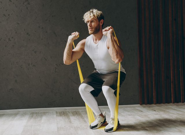 Full body side view young strong sporty athletic sportsman man wears white tank shirt black shorts using elastic rubber band for hands do squat warm up training indoor at gym. Workout sport concept