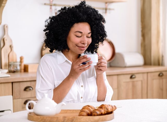 Smiling young african american woman sitting in kitchen and drinking her morning coffee with croissants. Tasty breakfast, concept of slow living and weekend rest at home. Positive beginning of new day