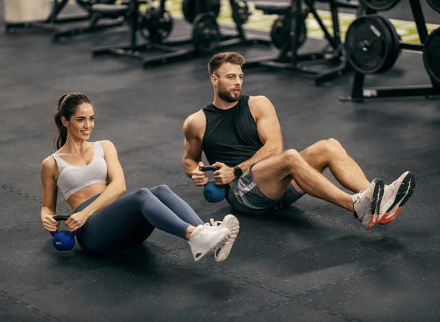 Fit muscular sportspeople doing russian twists with kettlebell on gym floor.