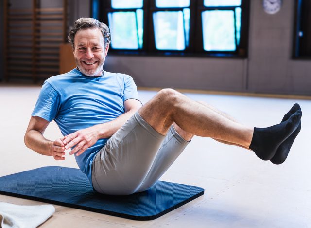 An active senior man is smiling while doing a sit up on a mat. He is wearing a blue shirt and gray shorts