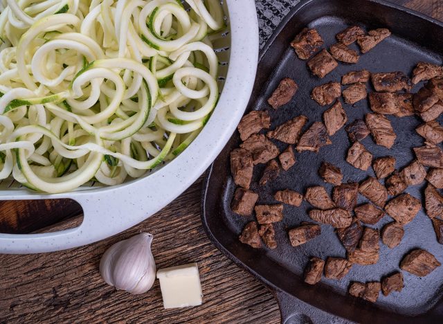 Raw, spiralized, zucchini noodles or zoodles in a colander with pan seared garlic steak bites, preparing to make garlic steak bites.