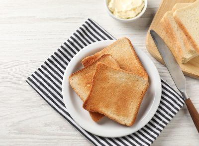 Delicious toasted bread slices with butter and knife on white wooden table, flat lay