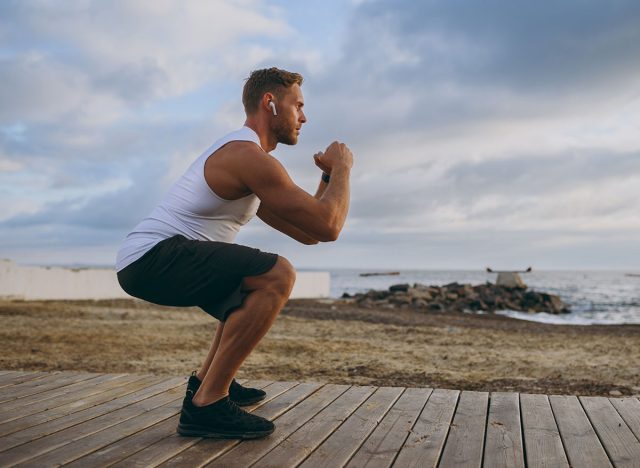 Full body young strong sporty athletic toned fit sportsman man wears sports clothes earphones do squats warm up train at sunrise sun dawn over sea beach outdoor on pier seaside in summer day morning