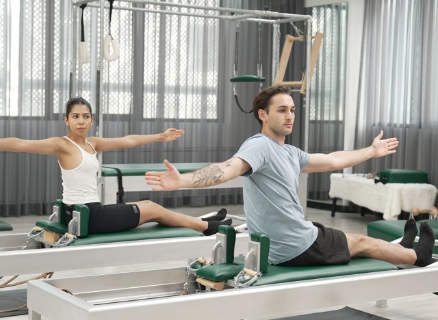 Two clients performing synchronized arm extensions on Pilates reformer machines. Happy pilates partner seated twist on the reformer for enhancing flexibility and building balance, control. Habituate.