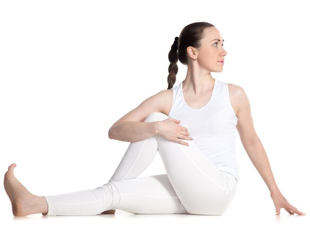 Sporty beautiful young woman in white sportswear practicing yoga, sitting in easy variation of Half lord of the fishes pose, vakrasana, studio full length isolated shot, front view