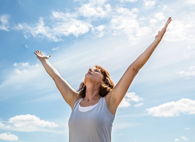 breathing outside - zen middle aged yoga woman looking up and raising her arms up,practicing meditation for freedom over summer blue sky,low angle view