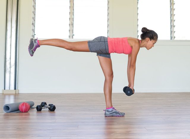 Woman strength training at gym exercising hamstring and lower back muscles with single-leg romanian deadlift exercises with free weights dumbbells. Asian girl alone indoors in fitness center room.