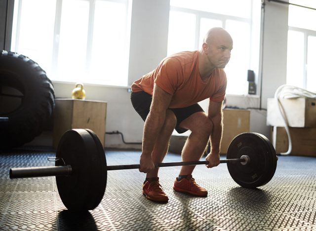 Middle-aged sporty man preparing to make exercise for biceps with help of barbell while having workout in modern gym