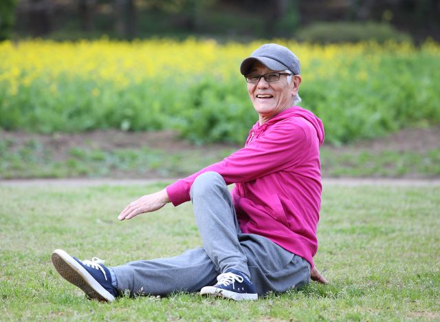 senior Japanese man wearing pink parka doing seated torso twist stretch on a lawn