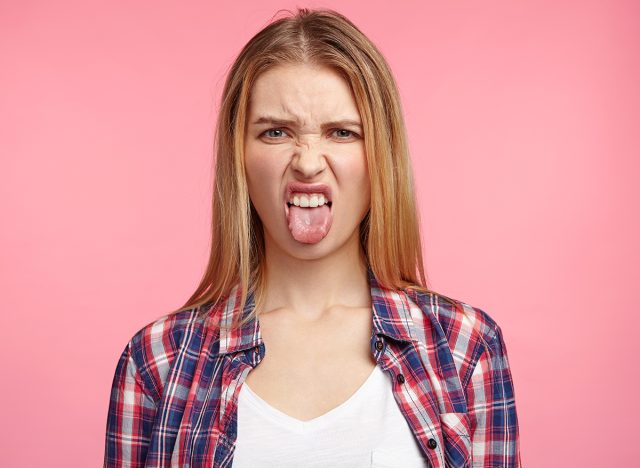 Dissatisfied female model frowns face, has disgusting expression, shows tongue, expresses non compliance, irritated with somebody, rejects do something. People and negative facial expressions, yuck