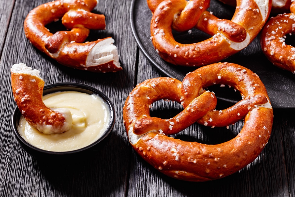 close-up of soft pretzels baked in the form of knot and sprinkled with salt on black plate on dark wooden table with cheese sauce, horizontal view from above